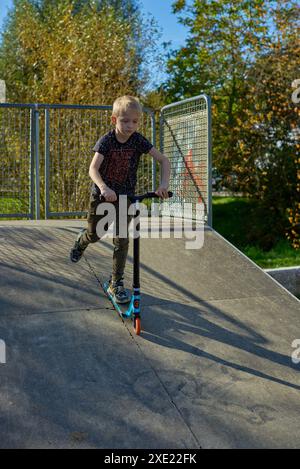 Junge auf dem Roller macht einen Trick und genießt seine Fahrt im Skatepark am bewölkten Frühlingstag. Junger Mann, der auf dem Kick sitzt Stockfoto