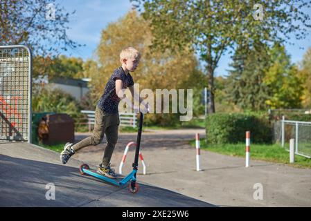 Junge auf dem Roller macht einen Trick und genießt seine Fahrt im Skatepark am bewölkten Frühlingstag. Junger Mann, der auf dem Kick sitzt Stockfoto