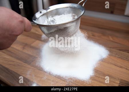 Eine Nahaufnahme einer männlichen Hand, die Mehl auf eine Holzfläche in einer Küche gesiebt hat Stockfoto