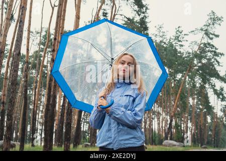 Junge Frau, die draußen im Wald einen transparenten blauen Regenschirm hält. Regenwetter mit Regenschirm. Frau mit Hand, die überprüft, wie Stockfoto