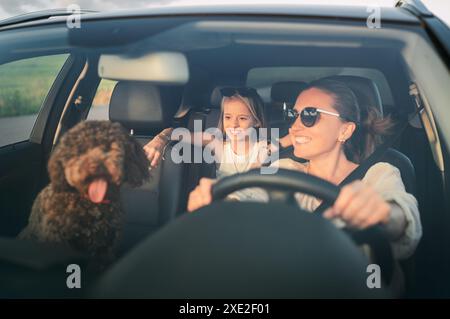 Lächelnde Frau, die Auto fährt, mit braunem Maltipoo-Hund und ihrer Tochter auf dem Rücksitz und genießt den sonnigen Tag. Das Bild zeigt einen fröhlichen Familienmoment während der Straße t Stockfoto