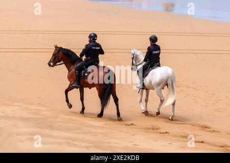 Gijon, Spanien - 24. Mai 2024: Zwei männliche spanische Polizeibeamte patrouillieren an einem sonnigen Tag zu Pferd einen Strand mit goldenem Sand Stockfoto