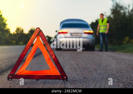 Nicht wiedererkennbarer trauriger Fahrer in reflektierender Weste. Männlicher Fahrer, der in der Nähe eines kaputten Autos steht, mit offener Motorhaube. Rotes Dreieck, um andere zu warnen Stockfoto