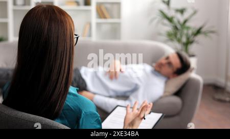 Ein junger Mann liegt auf einer Couch bei einem Psychologen-Termin. Eine brünette Brillenpsychologin führt eine psychologische Sitzung durch Stockfoto