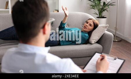 Eine junge Frau liegt auf einer Couch beim Termin eines Psychologen. Ein brauner, männlicher Psychologe mit Brille führt eine psychologische Sitzung durch Stockfoto