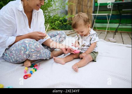 Eine Mutter und ihr Kleinkind teilen sich einen verspielten Moment auf einer Decke im Freien. Das Kind ist mit bunten Spielzeugen beschäftigt und die Mutter lächelt warm. Stockfoto