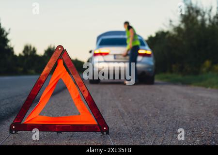 Nicht wiedererkennbarer trauriger Fahrer in reflektierender Weste. Männlicher Fahrer, der in der Nähe eines kaputten Autos steht, mit offener Motorhaube. Rotes Dreieck, um andere zu warnen Stockfoto