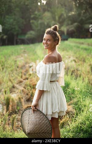 Glückliche junge Frau, die natürliche Kleidung trägt, die asiatischen konischen Hut auf dem Reisfeld hält Stockfoto