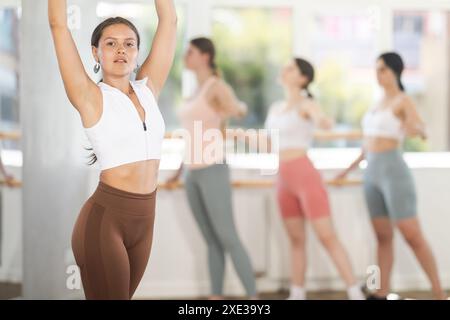 Porträt eines fröhlichen, aktiven Mädchens, das Ballettbewegungen im Fitnessstudio ausübt. Gruppe junger Balletttänzer im Hintergrund in der Nähe der Ballettbarre Stockfoto