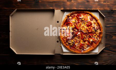 Peperoni Pizza mit Paprika und Zuckermais in offener Schachtel auf dunklem Holztisch flach gelegt mit Stockfoto