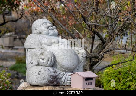 Nagahama, Präfektur Shiga, Japan. November 2023. Eine sitzende, lächelnde Buddha-Steinfigur an einem japanischen Shinto-Schrein. Stockfoto