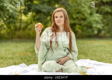 Weiße, hübsche schwangere Frau sitzt auf Decke, hält, isst Apfel im Park. Ruhe auf der Wiese, Entspannung. Gesunder Lebensstil. Die Entbindung Stockfoto