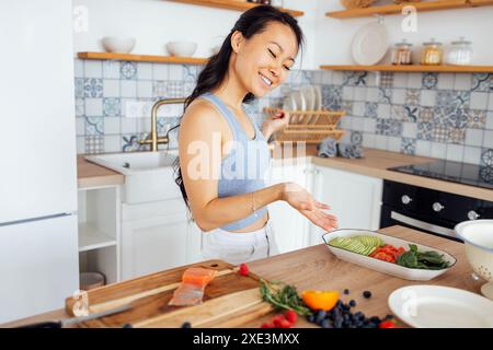 Eine attraktive süße asiatische Frau lächelt und kocht das Frühstück in der Küche. Eine junge charmante Koreanerin lacht und schneidet Vegetabl Stockfoto