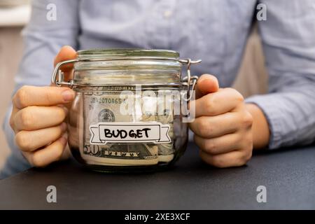 Unerkennbare Frau, die Spargeld in einem Glasgefäß hält, gefüllt mit Dollarscheine. BUDGET-Transkription vor dem Glas. Mana Stockfoto