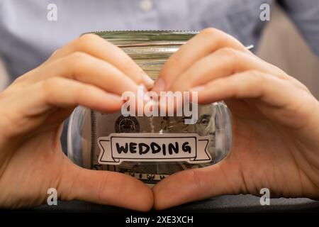 Unerkennbare Frau, die Herzzeichen zeigt, spart Geld in einem Glasgefäß, gefüllt mit Dollarscheine. Die UNKRAUTTRANSKRIPTION vor der Nase Stockfoto