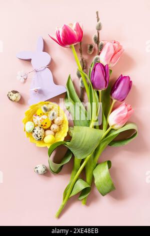 Rosa und lila Tulpen mit bunten Wachteleiern in einem Nest auf rosa Hintergrund. Frühjahrs- und Osterferienkonzept. Draufsicht flach Stockfoto