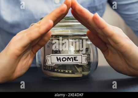 Unerkennbare Frau, die Spargeld in einem Glasgefäß hält, gefüllt mit Dollarscheine. HEIMTRANSKRIPTION vor dem Becher. Managi Stockfoto