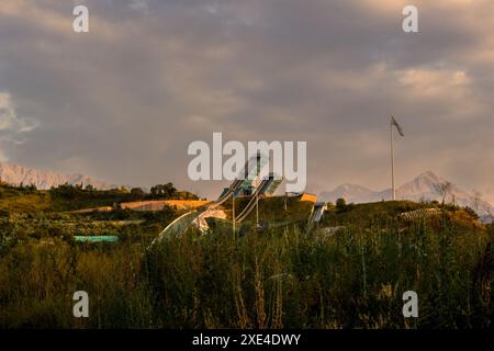 Der Sunkar International Skisprungkomplex mit bewölktem Abendhimmel und schneebedeckten Bergen im Hintergrund in Almaty, Kasachstan Stockfoto