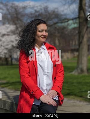 Romantisches Porträt einer stilvollen Frau in rotem Mantel und weißer Bluse. Positive Stimmung. Eine Dame läuft durch einen Stadtpark, sitzt auf der Attika und genießt einen Stockfoto