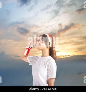 Patriotische junge Frau, die mit einer indonesischen Flagge auf der Wange grüßt, die ihren Nationalstolz mit einem wunderschönen Sonnenuntergangshimmel als Hintergrund zeigt Stockfoto