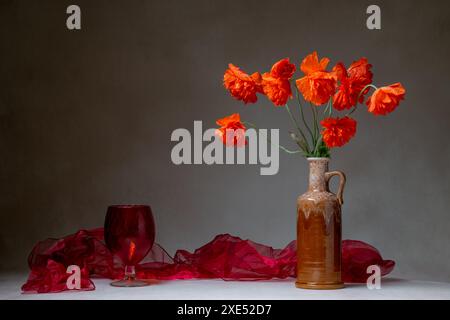 Ein Blumenstrauß aus rotem Mohn in einer hellbraunen Vase, neben einem roten Schal und einem roten Glas, isoliert auf hellem Hintergrund. Stillleben mit Platz für Text Stockfoto