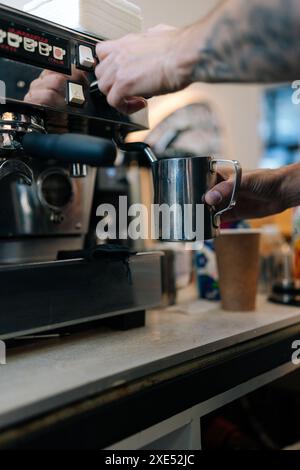 Vertikal geschnittene Aufnahme von nicht erkennbarer männlicher Barista, die Milch im aufschäumenden Pitcher aufschäumenden Kaffee mit einem professionellen Cappuccinator für die Kaffeemaschine im Café aufschäumenden Stockfoto