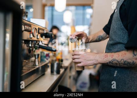 Zugeschnittene Aufnahme eines nicht erkennbaren Barista-Mannes, der leckeres Kaffeegetränk für Gäste in einem kleinen Business-Café zubereitete und einen Metalltopf hält, der Kaffee hineingießt Stockfoto