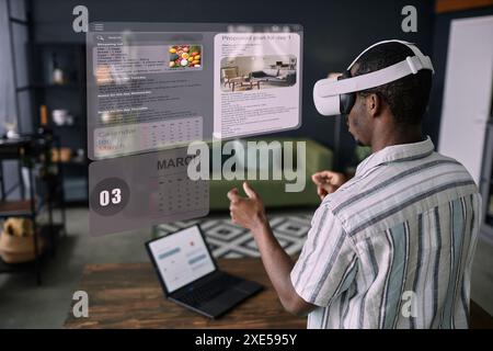 Junger schwarzer Mann mit VR-Headset, der im Wohnzimmer mit Zeitmanagement-Apps steht Stockfoto