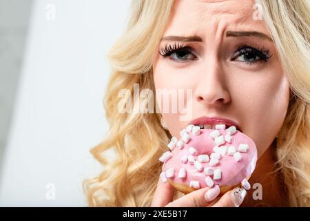 Die blonde Frau gibt sich einem schuldigen Vergnügen hin, sie beißt gierig in einen rosafarbenen, mit Marshmallows bedeckten Donut und befriedigt ihren süßen Zahn mit einem Zucker Stockfoto