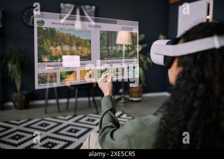 Überschulteraufnahme einer nicht erkennbaren Frau mit VR-Headset, die im Wohnzimmer sitzt und im Internet surft Stockfoto