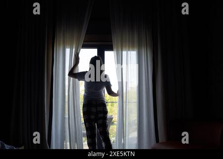 Frau, die Vorhänge öffnet, um das malerische Morgenlicht zu erstrahlen, steht an einem Fenster. Konzept, den Tag mit einer ruhigen Aussicht zu beginnen. Stockfoto