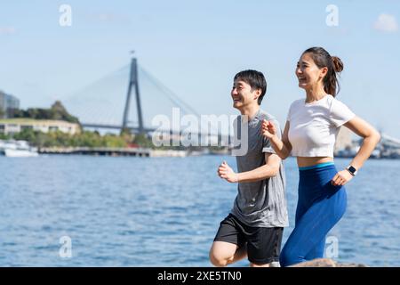 Männliche und weibliche Paare laufen mit einer Brücke im Hintergrund mit einem Lächeln im Gesicht Stockfoto