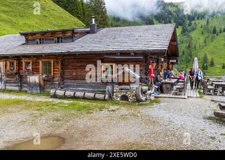Gäste sitzen auf der Terrasse einer Almhütte mit einem dunklen Ziegelfrontgiebel entlang des Salzburger Almenwegs bei Langbrand im Großarltal, umgeben von Nebelschwaden über den Bergen, Salzburg, Österreich. Eine rot-weiße Fahne und ein Sonnenschirm stehen am Eingang. Stockfoto