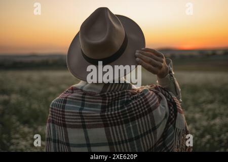 Eine Frau mit Hut und rot-weiß karierter Decke steht auf einem Feld. Die Sonne untergeht und strahlt ein warmes Leuchten über die Szene. Stockfoto