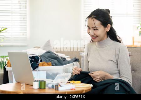 Frauen recyceln Kleidung zu Hause mit Nähzeug und Laptop, um nachhaltige Mode und umweltfreundliche Praktiken zu fördern Stockfoto