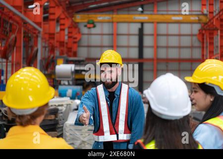 Vorgesetzter und Gruppe von Fabrikarbeitern, die Schutzhelme tragen, treffen sich kurz zusammen, bevor sie mit der Arbeit beginnen. Stockfoto