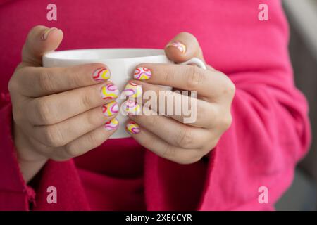 Pastellfarbene, weiche Nägel. Frau, die eine weiße Tasse Kaffee oder Tee hält, zeigt ihre neue Sommermaniküre in Farben von Stockfoto