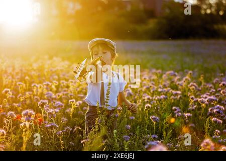 Wunderschönes Schulkind in einem Blumenfeld bei Sonnenuntergang, spielt mit Ariplane und Vintage Koffer, Frühling Stockfoto