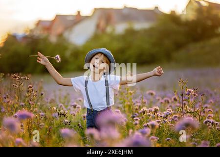 Wunderschönes Schulkind in einem Blumenfeld bei Sonnenuntergang, spielt mit Ariplane und Vintage Koffer, Frühling Stockfoto