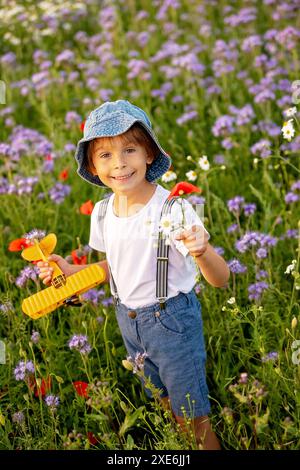 Wunderschönes Schulkind in einem Blumenfeld bei Sonnenuntergang, spielt mit Ariplane und Vintage Koffer, Frühling Stockfoto