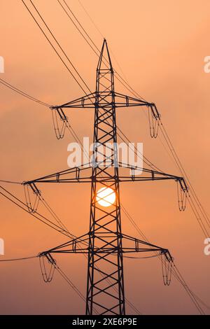 Untergehende Sonne hinter Silhouetten aus Strommasten und Stromleitungen, umhüllt von orangefarbenem Licht im Nebel. Kanton Zürich, Schweiz Stockfoto