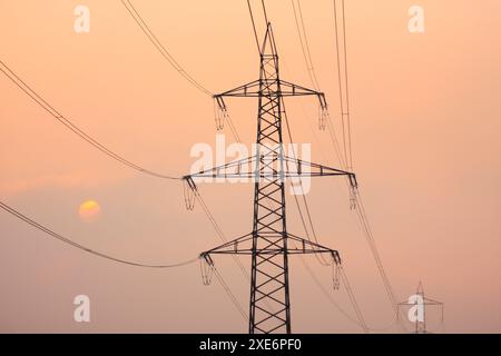 Untergehende Sonne hinter Silhouetten aus Strommasten und Stromleitungen, umhüllt von orangefarbenem Licht im Nebel. Kanton Zürich, Schweiz Stockfoto