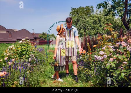 Glückliches junges Paar genießt Rosen Blumen, die im Sommergarten spazieren gehen. Gärtner kontrollieren Pflanzen mit Schürzen nach der Arbeit Stockfoto
