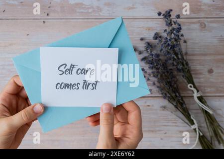 Weibliche Hände, die eine Papierkarte mit Text nehmen, KOMMEN ZUERST aus dem blauen Umschlag. Lavendelblume. Draufsicht, flach. Konz Stockfoto