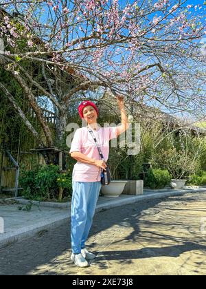 SA Pa Stadt, Lao Cai Provinz, Vietnam - 20. Februar 2024: Eine glückliche Touristenfrau neben Pfirsichblüten, die im Frühjahr in den hohen Bergen blüht Stockfoto