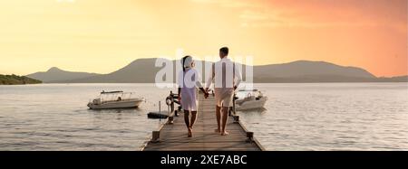 Pärchen, die bei Sonnenuntergang in Thailand auf einem hölzernen Pier im Meer spazieren gehen Stockfoto