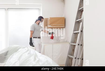 Mann Arbeiter, der das Produkt an die Wand spritzt, um Schimmel zu entfernen, Schutzhandschuhe und eine Atemschutzmaske mit einer Sprühflasche tragen. Sanierung der Schimmelwand b Stockfoto