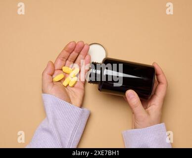 Die Hand einer Frau, die eine Plastikflasche und gelbe ovale Pillen auf braunem Hintergrund hält, von oben. Stockfoto