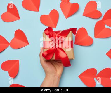Die Hand einer Frau hält eine Geschenkbox, die in ein rotes Seidenband auf blauem Hintergrund gewickelt ist Stockfoto