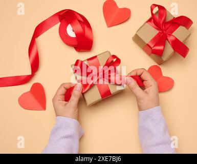 Die Hand einer Frau hält eine Geschenkbox in einem roten Seidenband auf beigefarbenem Hintergrund Stockfoto